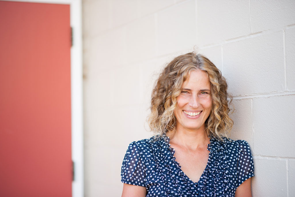 Gillian Jerome, leaning against a white brick wall, red door in the background. Smiling wearing a blue blouse with white dots.